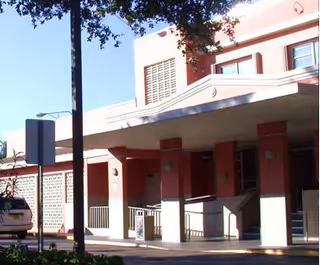 Exterior view of a building with a covered entrance supported by columns, a ramp with railings, and a tree partially visible in the foreground under a clear blue sky.