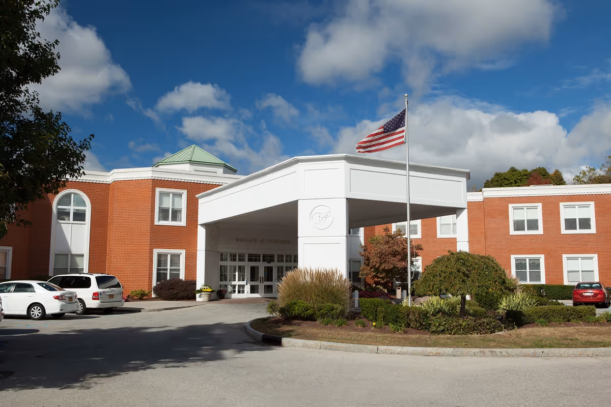 Exterior view of a brick senior living facility with a white covered entrance and an American flag on a flagpole in front. Several cars are parked near the entrance under a partly cloudy blue sky.