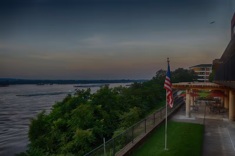 Riverside view from a terrace showing an American flag, patio seating under a pergola, and trees along the river at sunset.