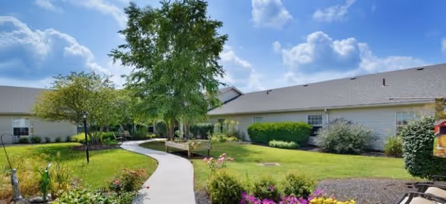 A sunny outdoor garden area with a curved concrete pathway, green grass, colorful flowers, and several trees. The pathway leads to a single-story building with beige siding and a gray roof under a blue sky with scattered clouds.