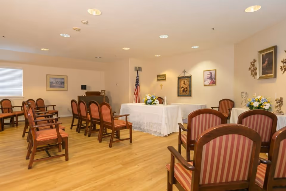 A small chapel or prayer room with wooden chairs arranged in rows facing a table covered with a white cloth. The table has floral arrangements and religious items on it. The walls are decorated with framed religious artwork and an American flag stands in the corner. The room has wooden flooring and recessed ceiling lights.