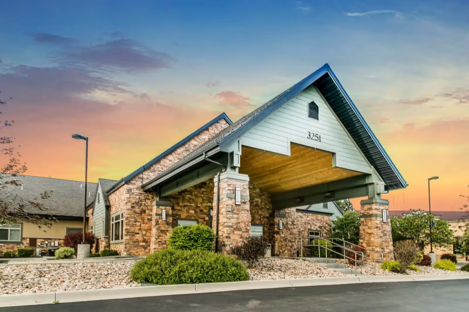 Exterior view of a senior living facility building with stone and wood siding, a covered entrance with the number 3251 displayed, surrounded by landscaping and a paved driveway under a colorful sunset sky.