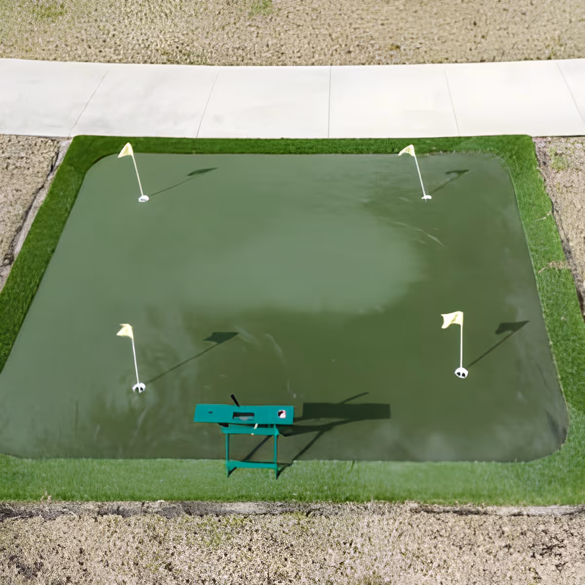 A small rectangular putting green with four yellow flags marking holes, surrounded by a narrow strip of green grass and bordered by gravel and a concrete walkway. A green golf ball return stand is positioned at one end of the putting green.