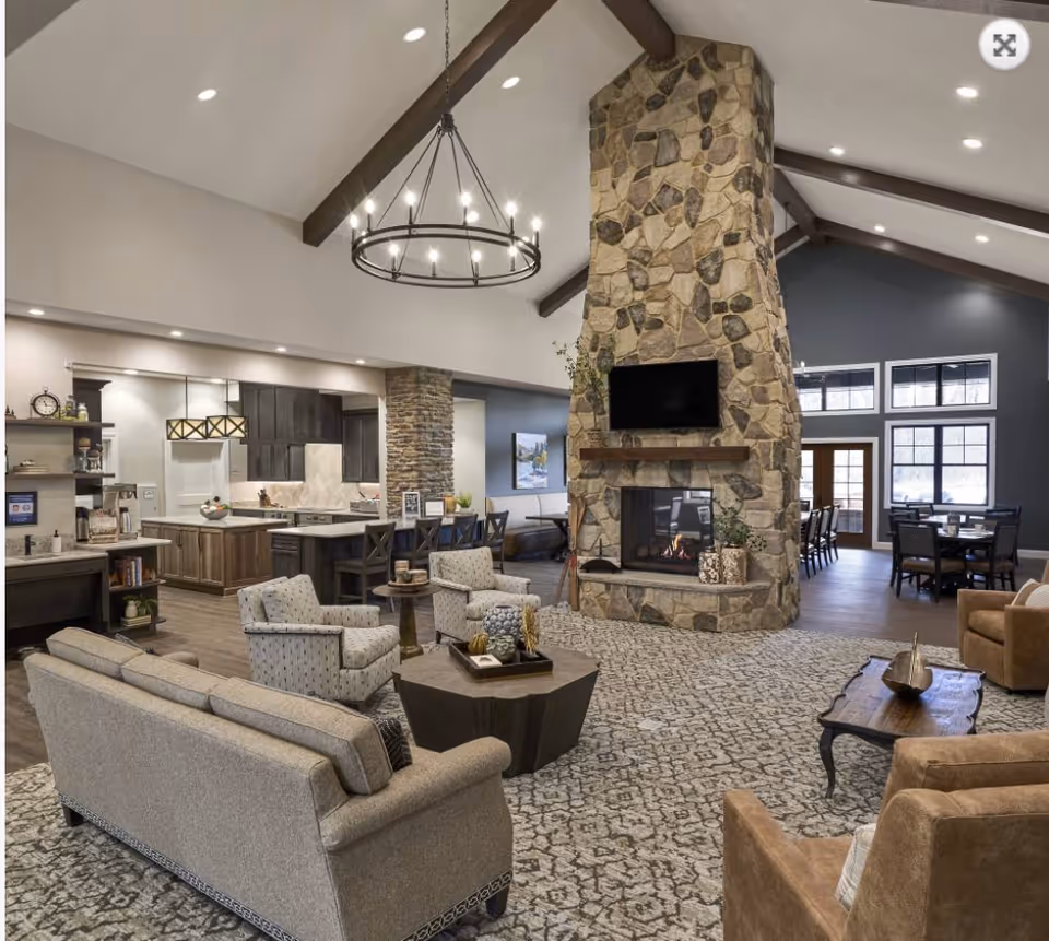 Spacious senior living common area featuring a large stone fireplace with a mounted TV above it, surrounded by comfortable seating including sofas and armchairs. The room has high vaulted ceilings with exposed wooden beams and a circular chandelier. In the background, there is an open kitchen with dark wood cabinets and a dining area with tables and chairs near large windows letting in natural light.