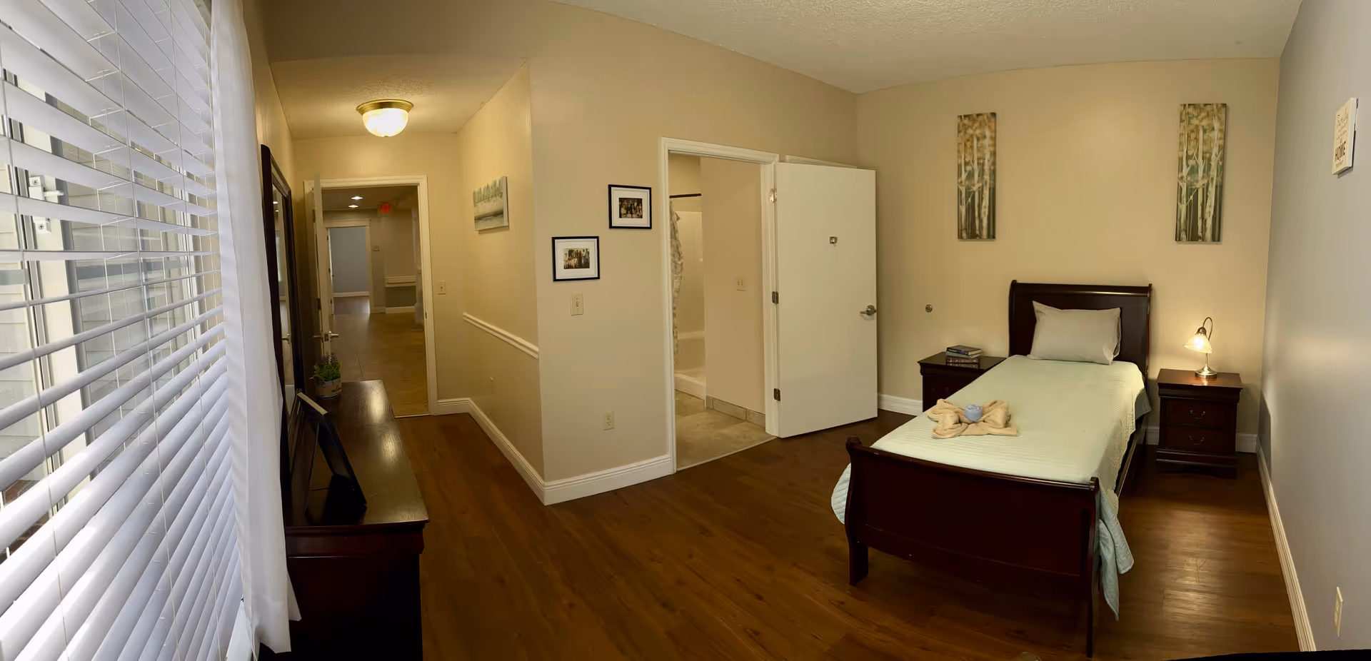A single bedroom in a senior living facility with a wooden bed frame, a neatly made bed with a light green bedspread and a pillow, two wooden nightstands with a lamp and books, two vertical paintings on the wall, and an open door leading to a bathroom. The room has wooden flooring and beige walls. A window with white blinds and a sheer curtain is on the left side, and a hallway is visible beyond the bedroom door.