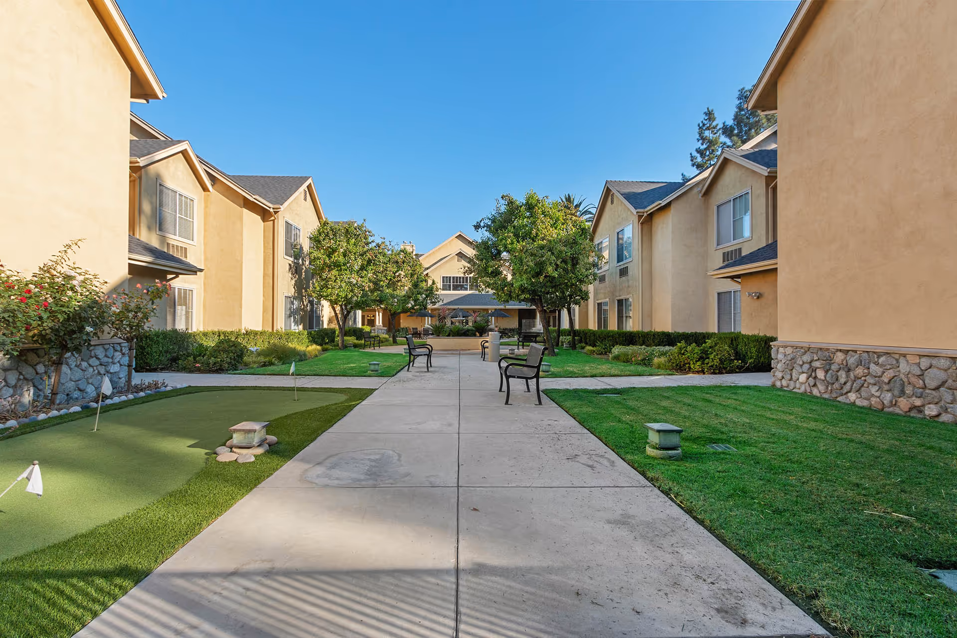 Outdoor courtyard area at Claremont Place featuring a paved walkway with benches on either side, green grass, trees, and a small putting green. Beige two-story buildings with stone accents surround the courtyard under a clear blue sky.