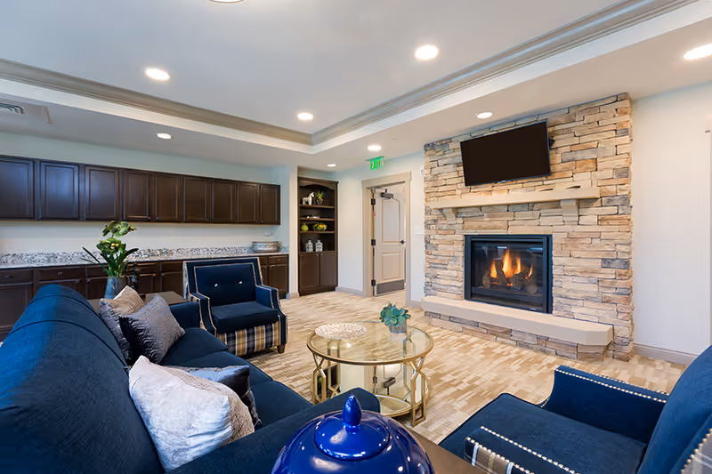 A cozy living room area with navy blue upholstered sofas and armchairs arranged around a glass coffee table. The room features a stone fireplace with a flat-screen TV mounted above it. There are dark wooden cabinets and shelves along one wall, decorated with plants and vases. The room is well-lit with recessed ceiling lights and has a beige patterned carpet.