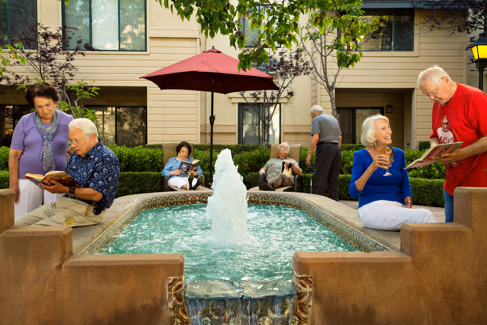 A group of older adults socializing and reading around a courtyard fountain outside a senior living building.