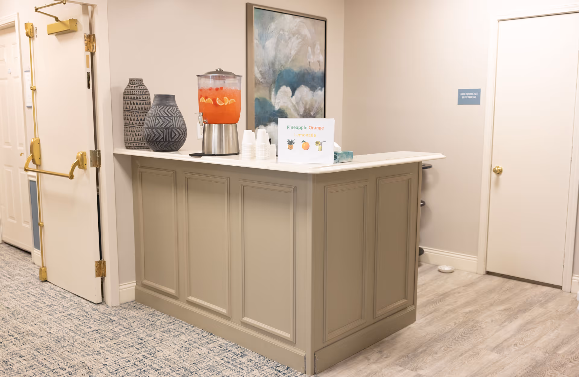 A beige reception or serving counter with a beverage dispenser filled with pineapple orange lemonade, two decorative vases, a stack of white cups, and a sign indicating the drink flavor. The area has light-colored walls, a carpeted floor, and two closed doors in the background.