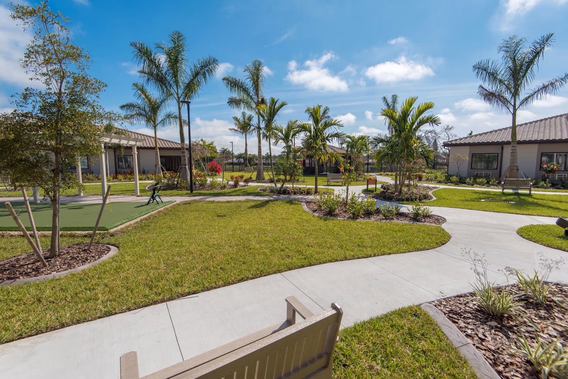 A sunny outdoor garden area with palm trees, green grass, paved walkways, benches, and small buildings with tiled roofs under a blue sky with scattered clouds.