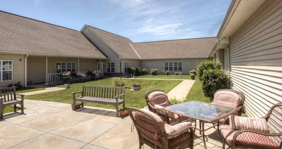 Outdoor courtyard area at Vintage Park at Ottawa featuring a paved patio with a glass-top table and four cushioned wicker chairs, wooden benches, green grass, potted plants, and a building with beige siding and multiple windows under a clear blue sky.