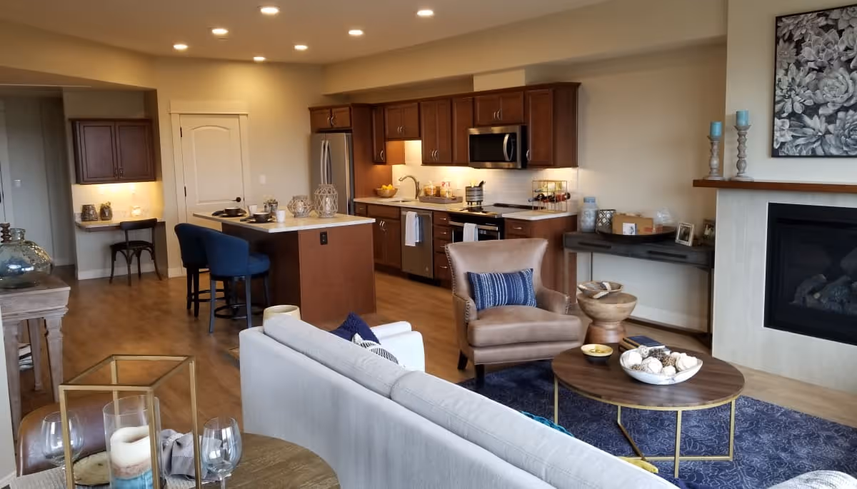 Open concept living area featuring a light gray sofa, a brown armchair with a blue pillow, a round wooden coffee table with decorative items, a fireplace with candles and a floral artwork above it, and a kitchen with wooden cabinets, stainless steel appliances, an island with two blue bar stools, and a small desk area with a chair.