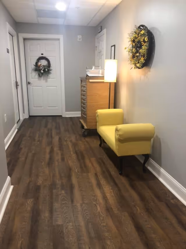 A well-lit interior hallway with wood flooring featuring a yellow upholstered bench, a floor lamp, a decorative wall wreath, and a closed white door.