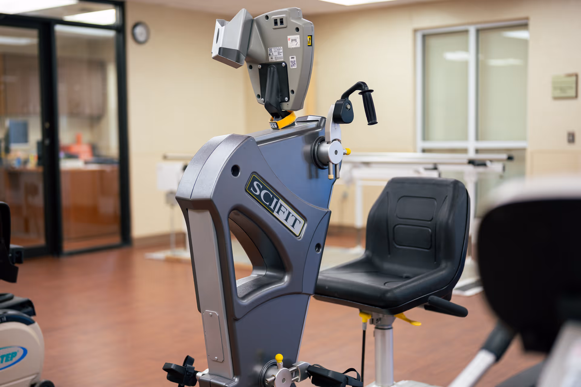 A close-up view of a SciFit exercise machine with a black seat in a rehabilitation or fitness room with wooden flooring and beige walls. In the background, there are parallel bars and a glass door leading to another room.
