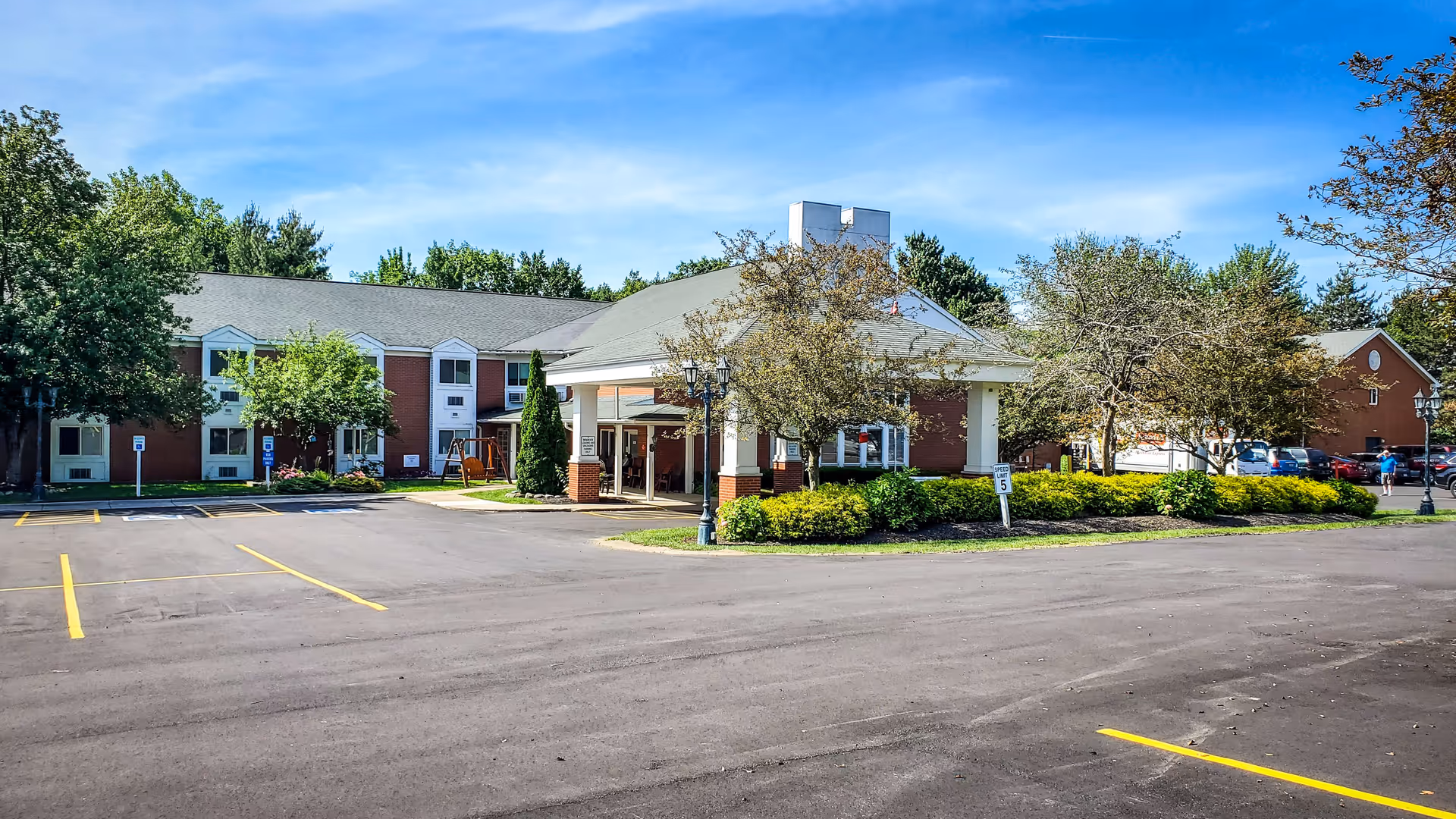 Front exterior of a brick senior living facility with a covered entrance, parking lot, and landscaped grounds under a blue sky.