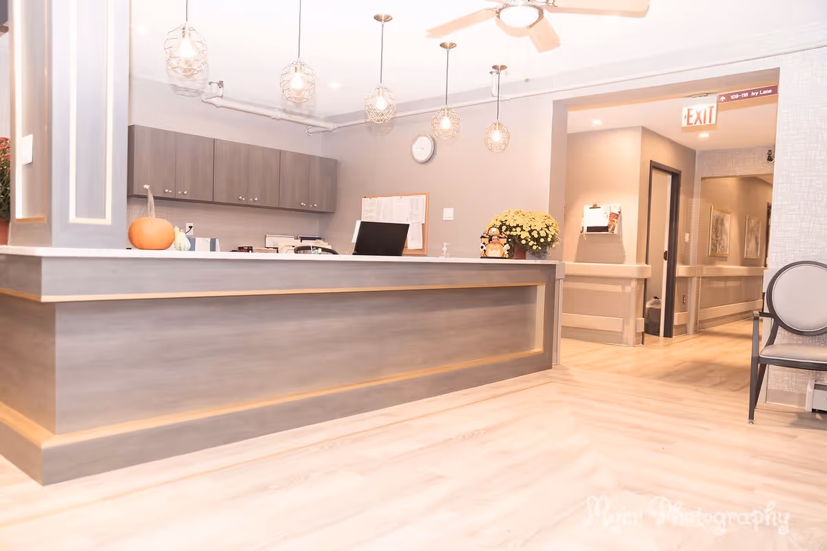 Reception area with a modern wooden front desk, pendant lights hanging from the ceiling, a clock on the wall, and a hallway leading to other rooms. There is a pumpkin and a flower pot on the desk, and a chair is partially visible on the right side.