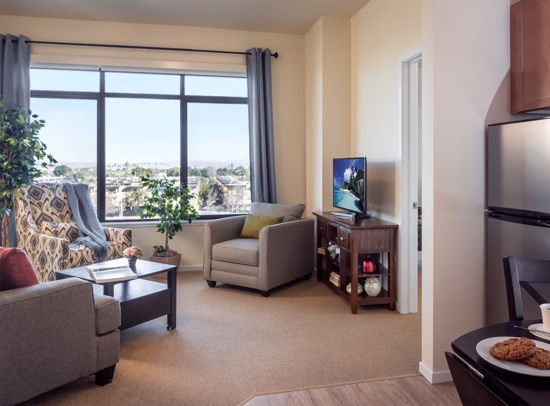 A cozy living room area with a large window showing an outdoor view. The room has a patterned armchair with a gray throw blanket, a gray armchair with a green pillow, a wooden coffee table with a book and a small plant, and a wooden TV stand with a flat-screen TV. There are two potted plants near the window, beige walls, and carpeted flooring. Part of a kitchen area with a stainless steel refrigerator and a table with cookies on a plate is visible on the right side.