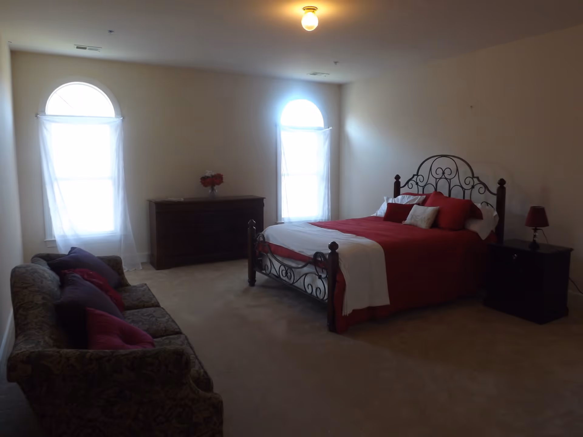 Spacious bedroom with an ornate metal bed dressed in red bedding, a couch, dresser, and two arched windows with sheer curtains.