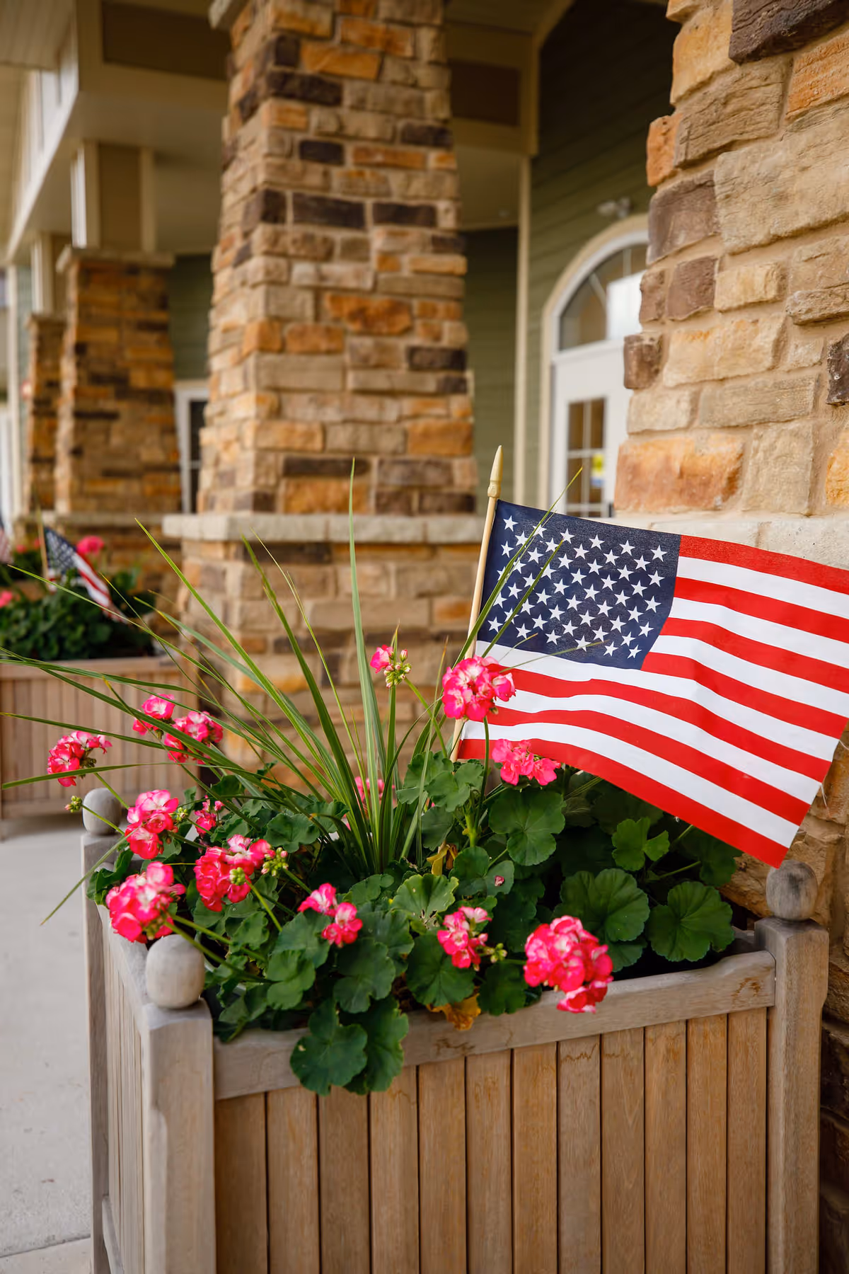 A wooden planter box filled with green plants and bright pink flowers, decorated with a small American flag. The planter is placed on a porch with stone pillars and a green exterior wall with a white door in the background.