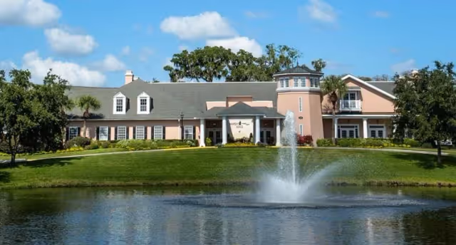 Exterior view of Marsh's Edge facility showing a large building with a gray roof and light pink walls, surrounded by trees and greenery. In front of the building is a pond with a fountain spraying water upwards under a blue sky with some clouds.