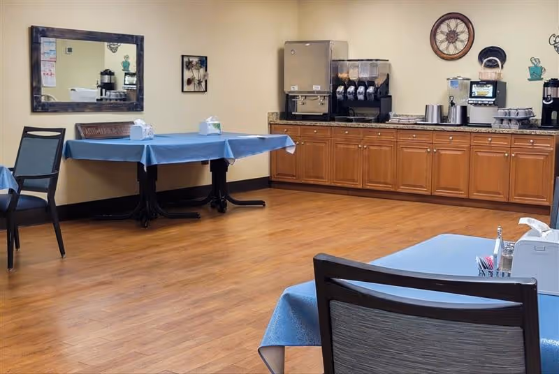 Community dining area with tables covered in blue tablecloths, chairs, and a countertop beverage station along the wall.