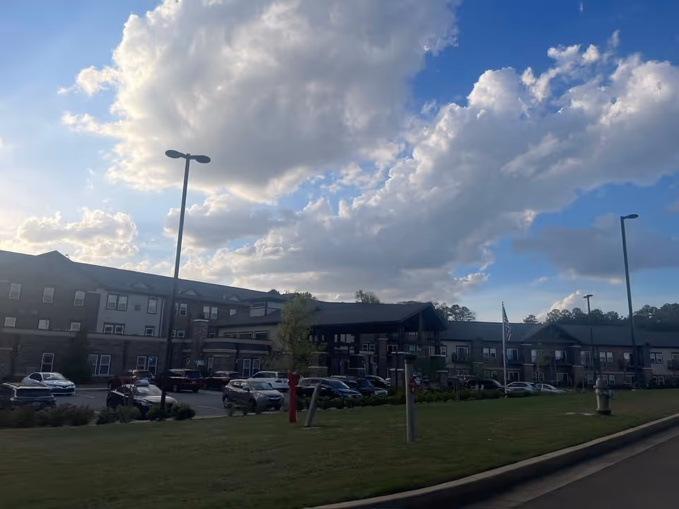 Front exterior of a multi-story senior living building with a parking lot, flagpole, and large clouds in the sky.