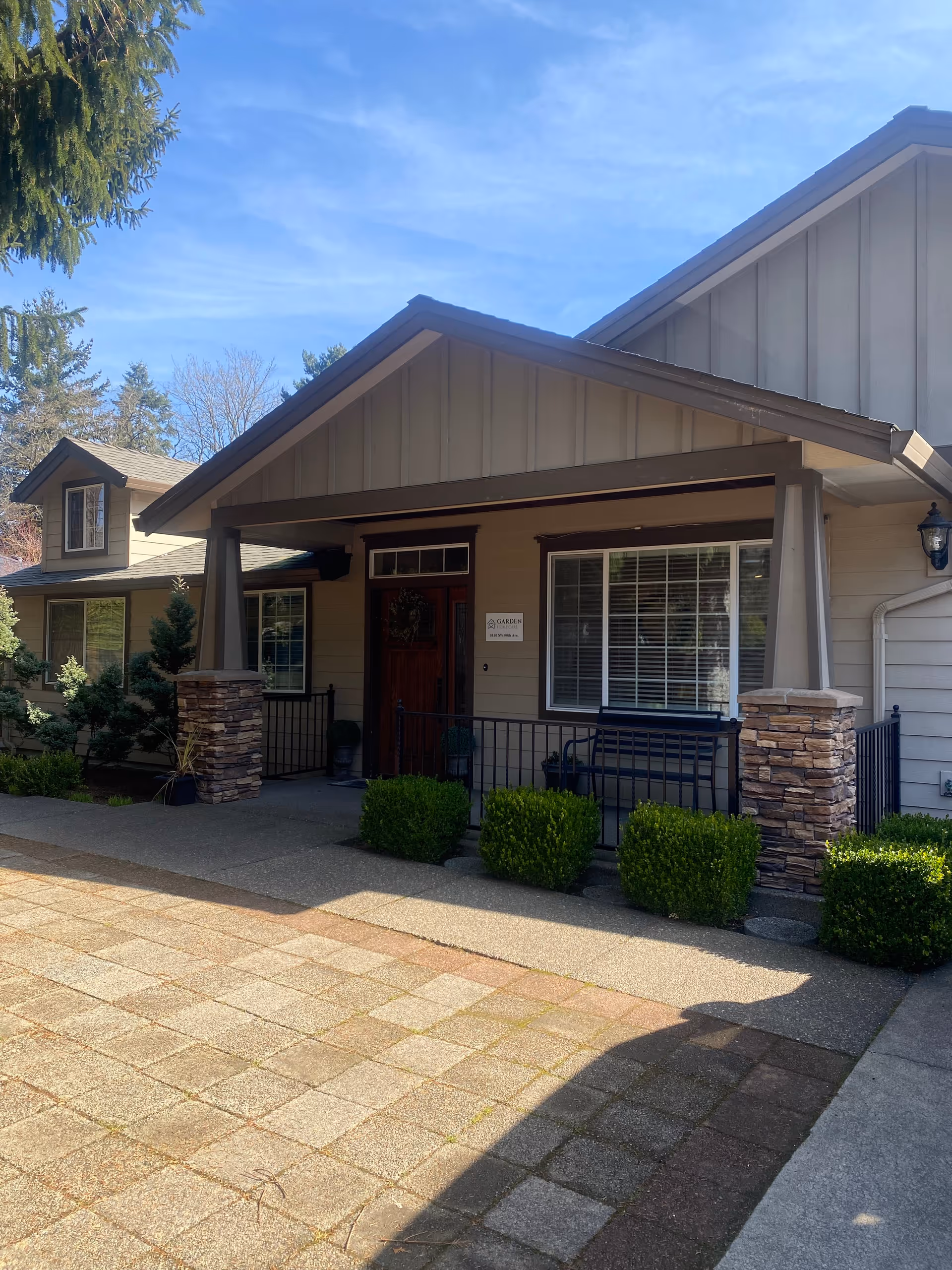 Exterior view of a single-story building with a covered porch supported by stone pillars. The building has beige siding with brown trim, a wooden front door, and a large window with white blinds. There are neatly trimmed bushes along the front and a paved walkway leading to the entrance. The sky is clear and blue.