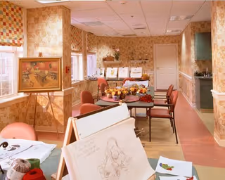 A cozy interior room with floral wallpaper and multiple tables and chairs arranged for activities. There are art supplies and sketches on one table in the foreground, and a framed painting on an easel near the windows. The room is well-lit with natural light coming through the windows.