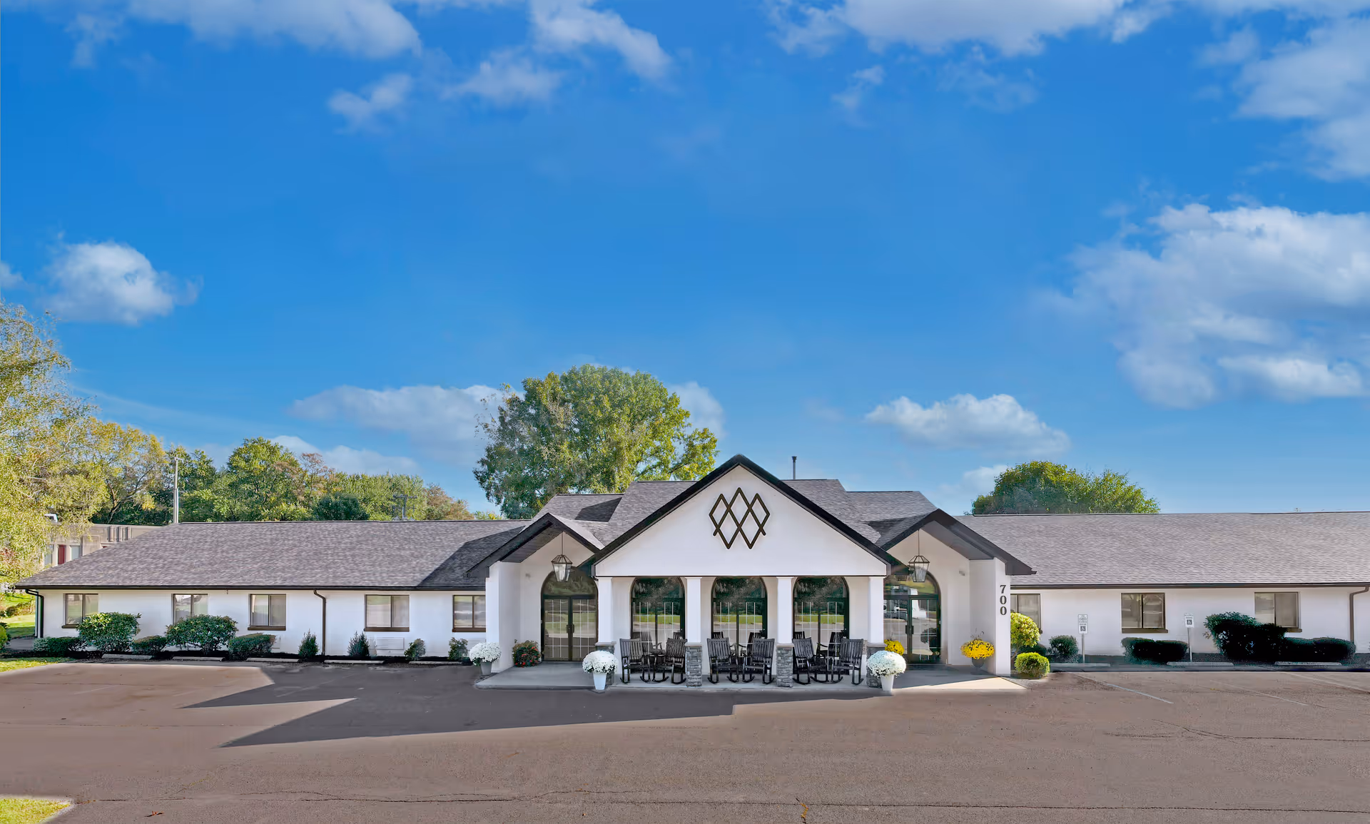 Front exterior view of a single-story senior living facility building with a covered entrance featuring large windows and outdoor seating. The building is white with a dark roof and surrounded by greenery under a blue sky with some clouds.