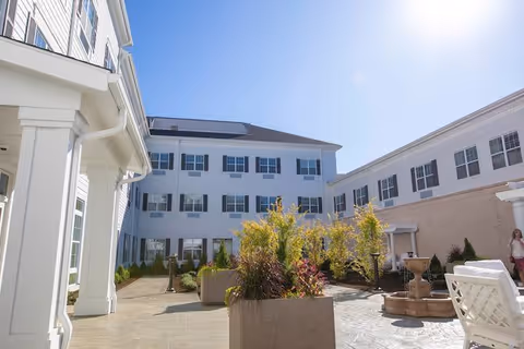 Sunny courtyard between white multi-story residential buildings with planters, seating, and a fountain.
