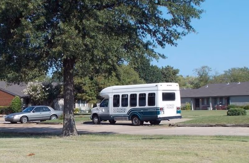 A white shuttle bus parked on a driveway in front of single-story residential buildings with a large tree and a silver car nearby under a clear blue sky.