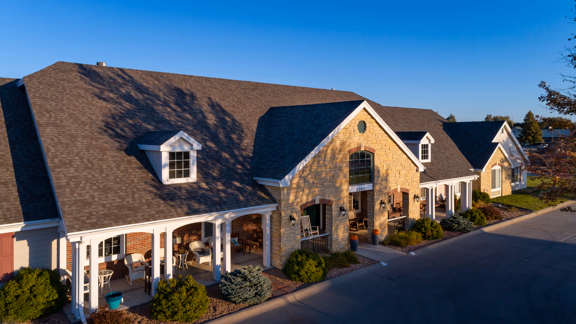 Exterior view of a single-story assisted living facility building with a combination of stone and brick facade, multiple windows, and a covered porch area with chairs. The building is surrounded by small bushes and landscaping under a clear blue sky.