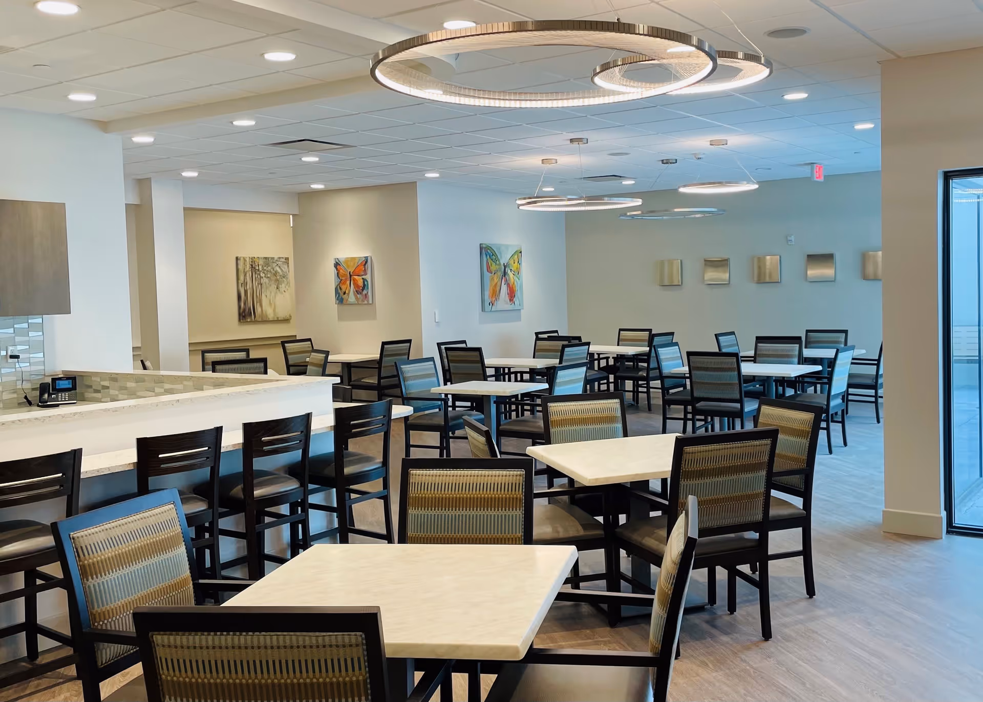 A modern dining area with multiple square tables and chairs arranged neatly. There is a counter with high chairs on the left side. The room is well-lit with circular ceiling lights and decorated with colorful butterfly paintings on the walls. Large windows on the right allow natural light to enter.
