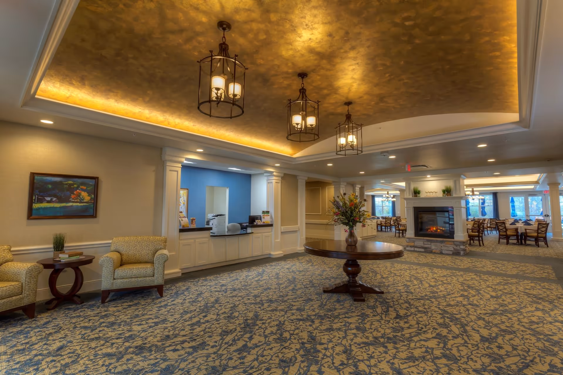 Spacious, elegant senior living interior with seating, a central round table, fireplace, chandeliers and a dining area in the background.