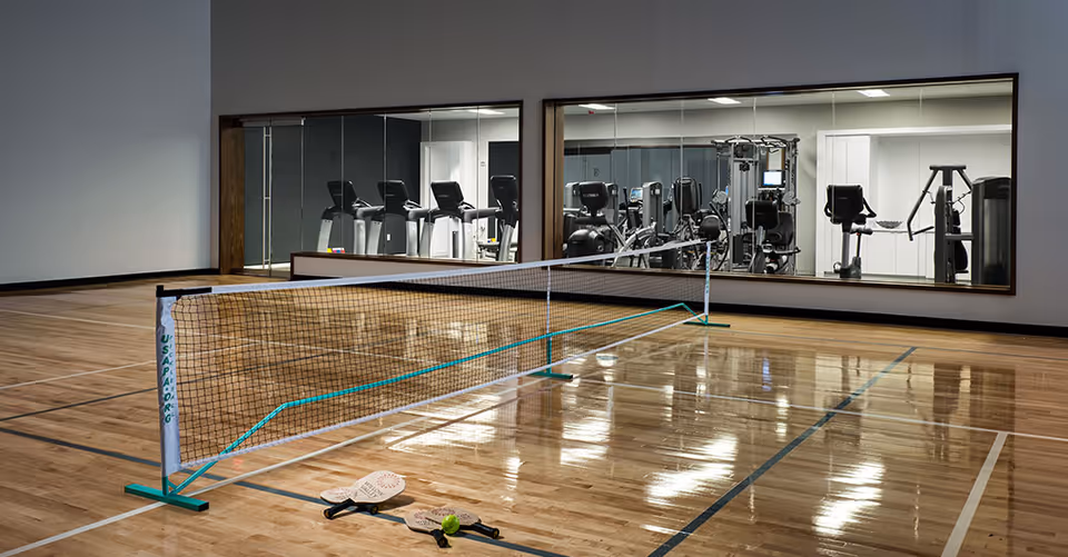Indoor gymnasium with a pickleball net set up on a polished wooden floor. Two pickleball paddles and a ball are placed on the floor near the net. In the background, there is a large window showing a fitness room with various exercise machines including treadmills and weight equipment.