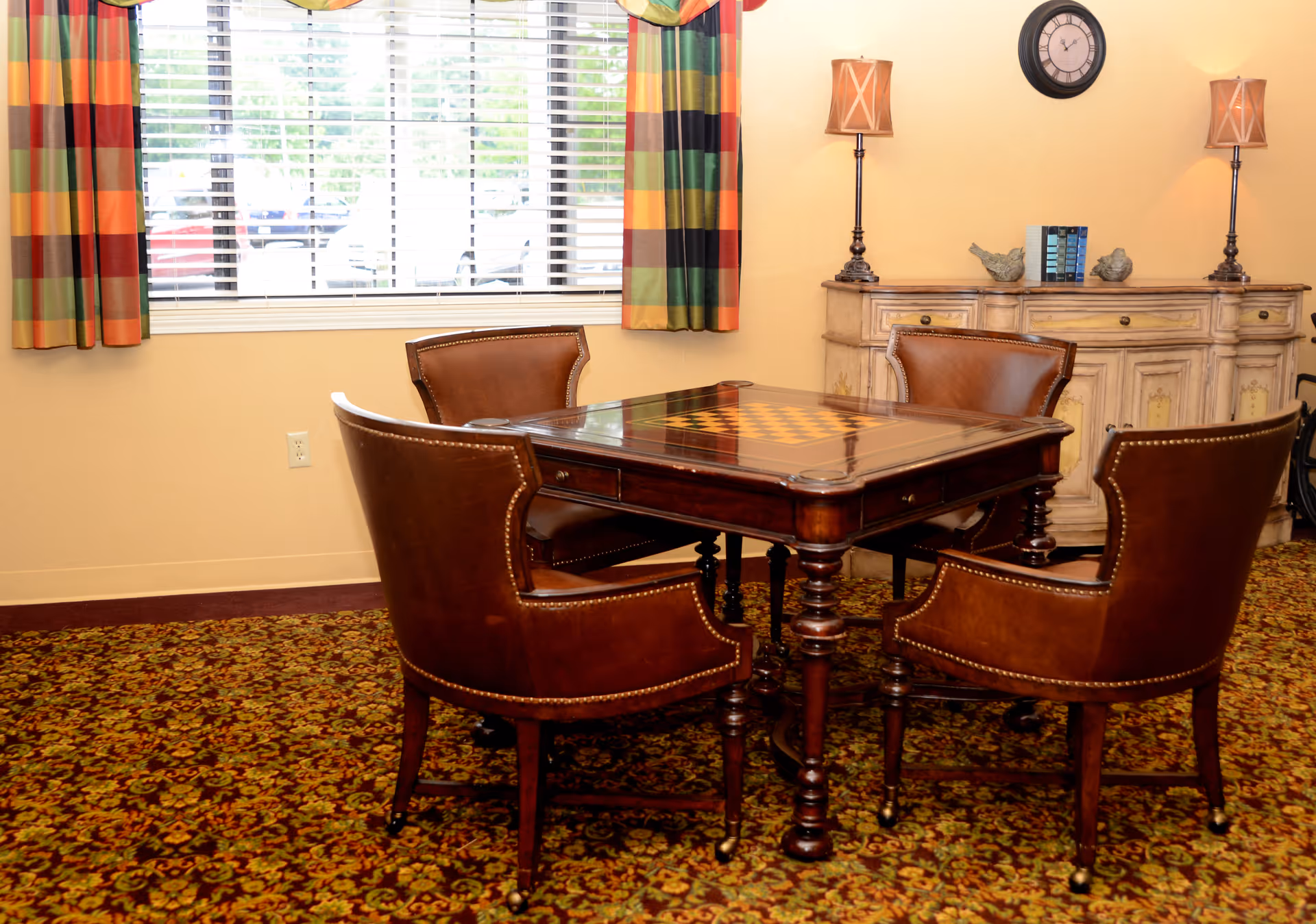 A cozy interior room with a square wooden table featuring a chessboard pattern on top, surrounded by four brown leather chairs. Behind the table is a cream-colored sideboard with two lamps and decorative items on it. A window with colorful plaid curtains lets in natural light, and a clock hangs on the wall above the sideboard. The floor is covered with a patterned carpet.