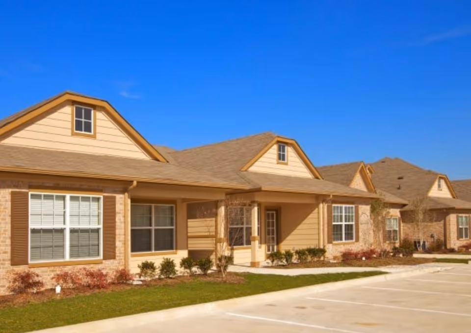 Exterior view of a single-story residential building with beige siding, brown shutters, and a sloped roof under a clear blue sky. There are small bushes and landscaping in front of the building and a paved parking area in the foreground.