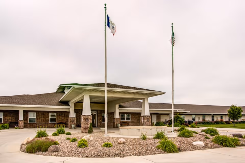 Exterior view of a single-story brick building with a covered entrance supported by white pillars. Two flagpoles with flags stand in front of the entrance, surrounded by landscaped rocks and greenery. The sky is overcast.