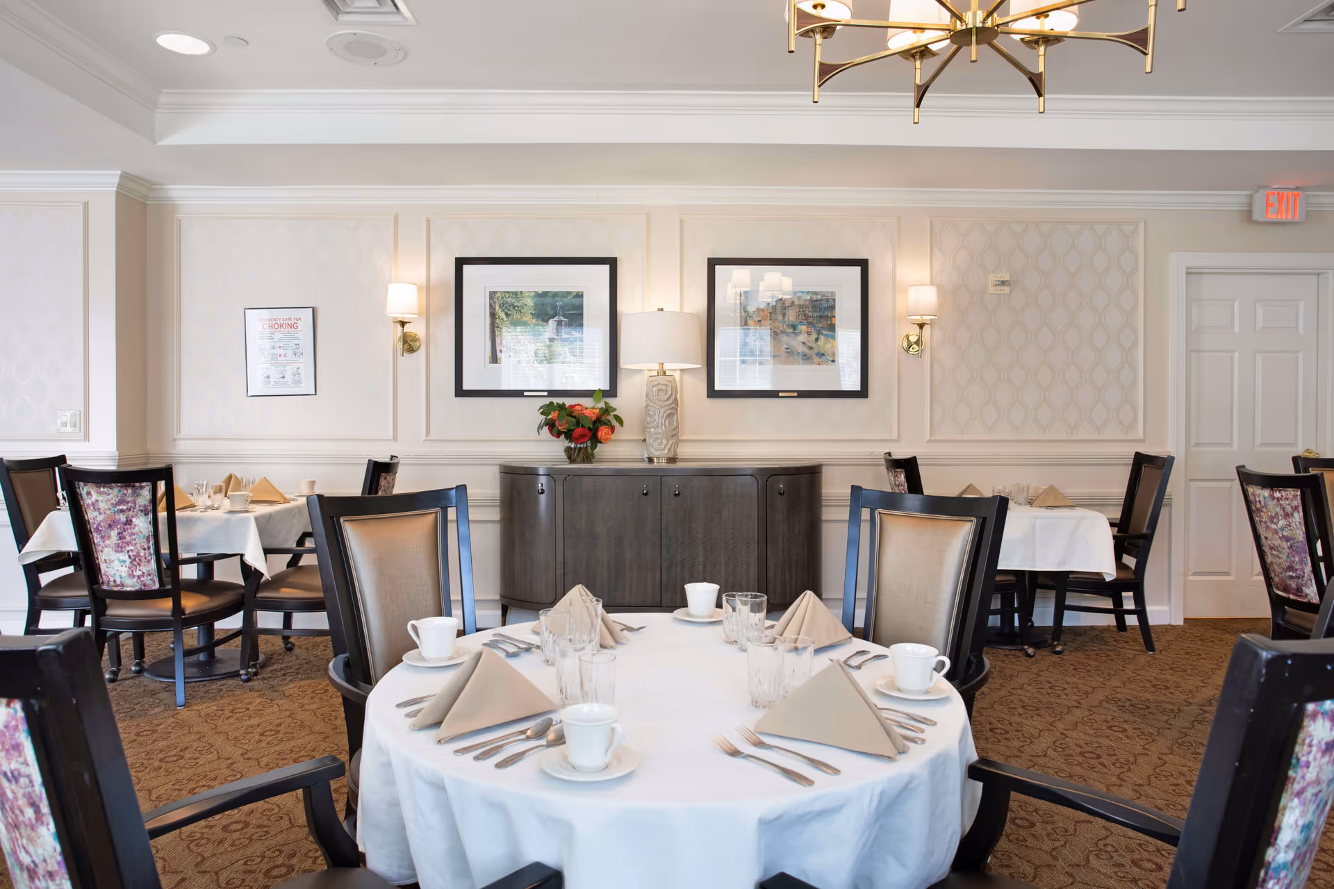A dining room in a senior living facility with round tables covered in white tablecloths, set with cups, glasses, napkins, and silverware. The room has upholstered chairs, patterned carpet, framed artwork on the walls, wall sconces, a sideboard with a lamp and flowers, and a chandelier hanging from the ceiling.