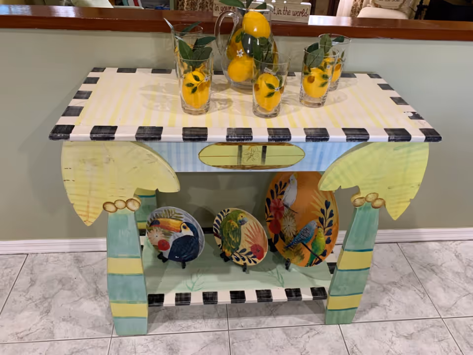 A decorative wooden table painted with pastel colors and black-and-white checkered edges. On the tabletop, there is a pitcher and three glasses filled with artificial lemons and green leaves. Below the table, there are three decorative plates featuring colorful bird illustrations. The table is placed on a tiled floor against a light green wall.