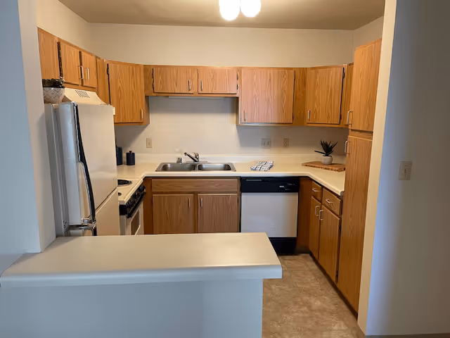 A compact kitchen in a senior living facility featuring wooden cabinets, a white refrigerator, a stove with an oven, a double sink, a dishwasher, and a small countertop bar area. The kitchen has beige walls and a tiled floor.