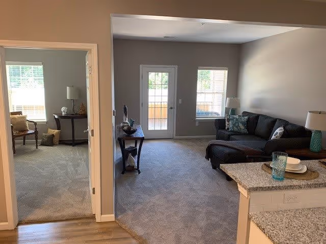 Open-plan living area with a sectional sofa and ottoman, console table, glass door to a balcony, and a partial view of a kitchen counter and adjacent room.