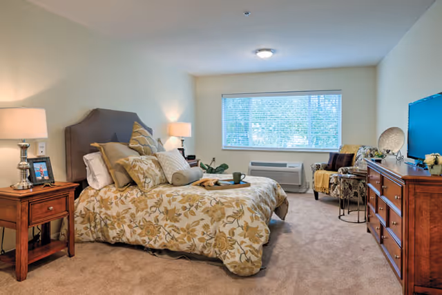 A cozy bedroom with a large bed covered in floral patterned bedding and multiple pillows. There are two wooden nightstands with lamps on either side of the bed. A window with blinds lets in natural light, and an air conditioning unit is installed below the window. Across from the bed is a wooden dresser with a flat-screen TV on top. A comfortable armchair with a throw blanket and a small side table are placed near the window.