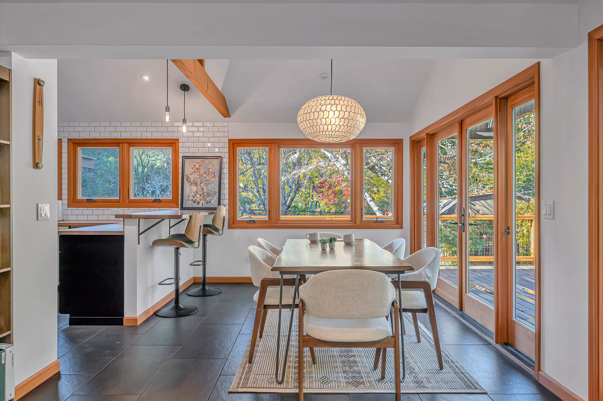 Bright dining area with a wooden table and upholstered chairs, pendant light, large windows and glass doors opening to a deck, and a small counter with bar stools.