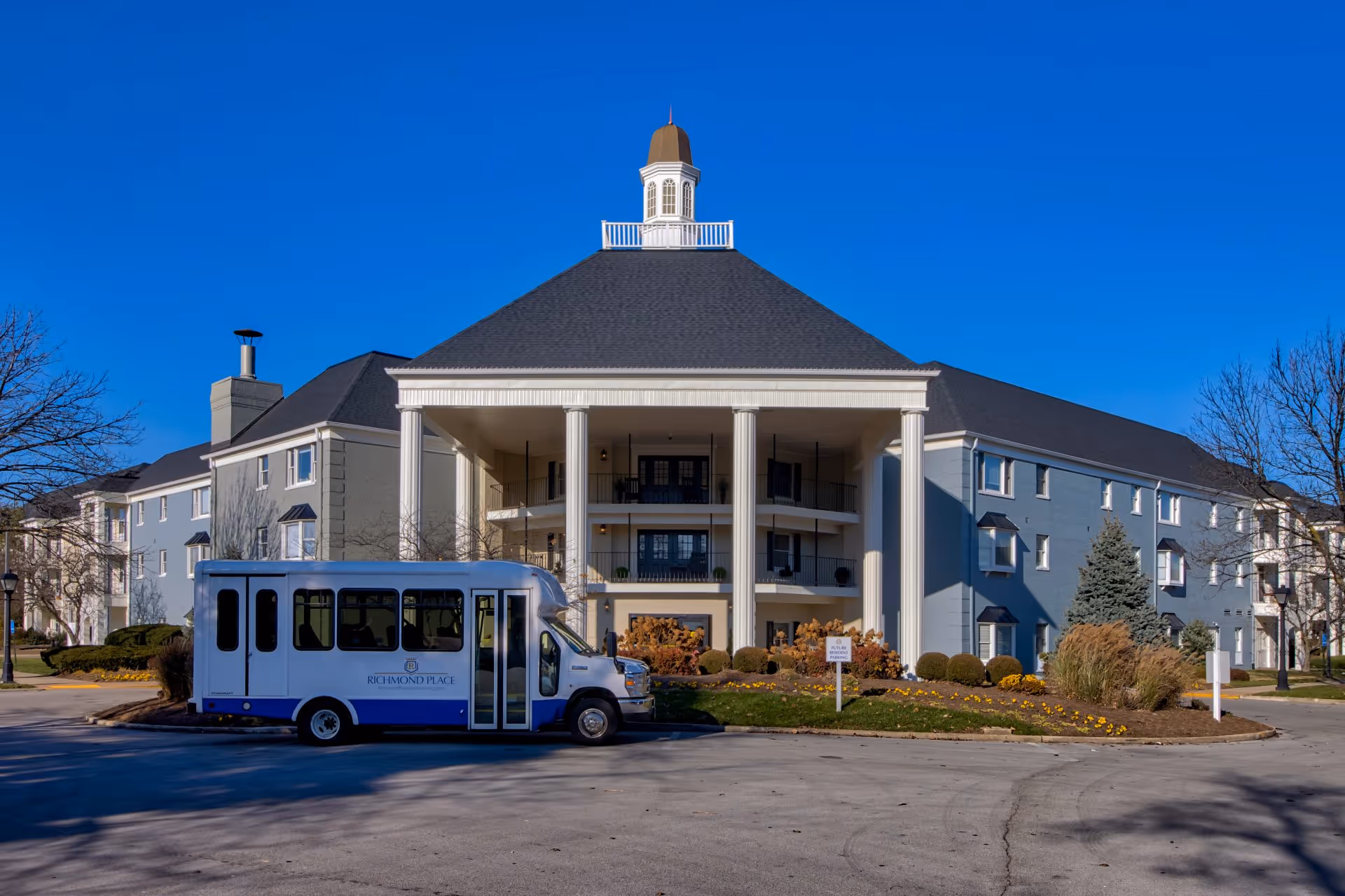 Exterior view of Richmond Place senior living facility showing a large building with a prominent central section featuring tall white columns and a cupola on the roof. A white and blue shuttle bus with Richmond Place branding is parked in front of the building. The sky is clear and blue, and there are landscaped bushes and flowers around the entrance.