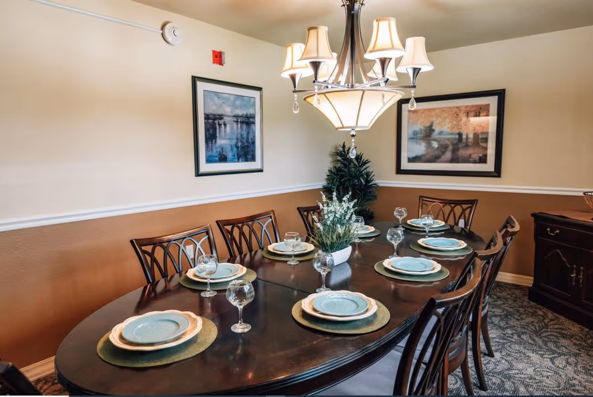Formal dining room with a dark wood table set for eight, plates and glassware, a chandelier overhead and framed artwork on the walls.