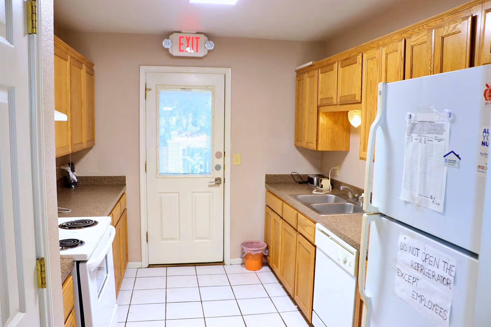 A clean kitchen with wooden cabinets on both sides, a white stove on the left, a white refrigerator on the right with a sign that reads 'DO NOT OPEN THE REFRIGERATOR EXCEPT EMPLOYEES,' a dishwasher, a double sink, and a door with a window and an illuminated exit sign above it at the end of the room.