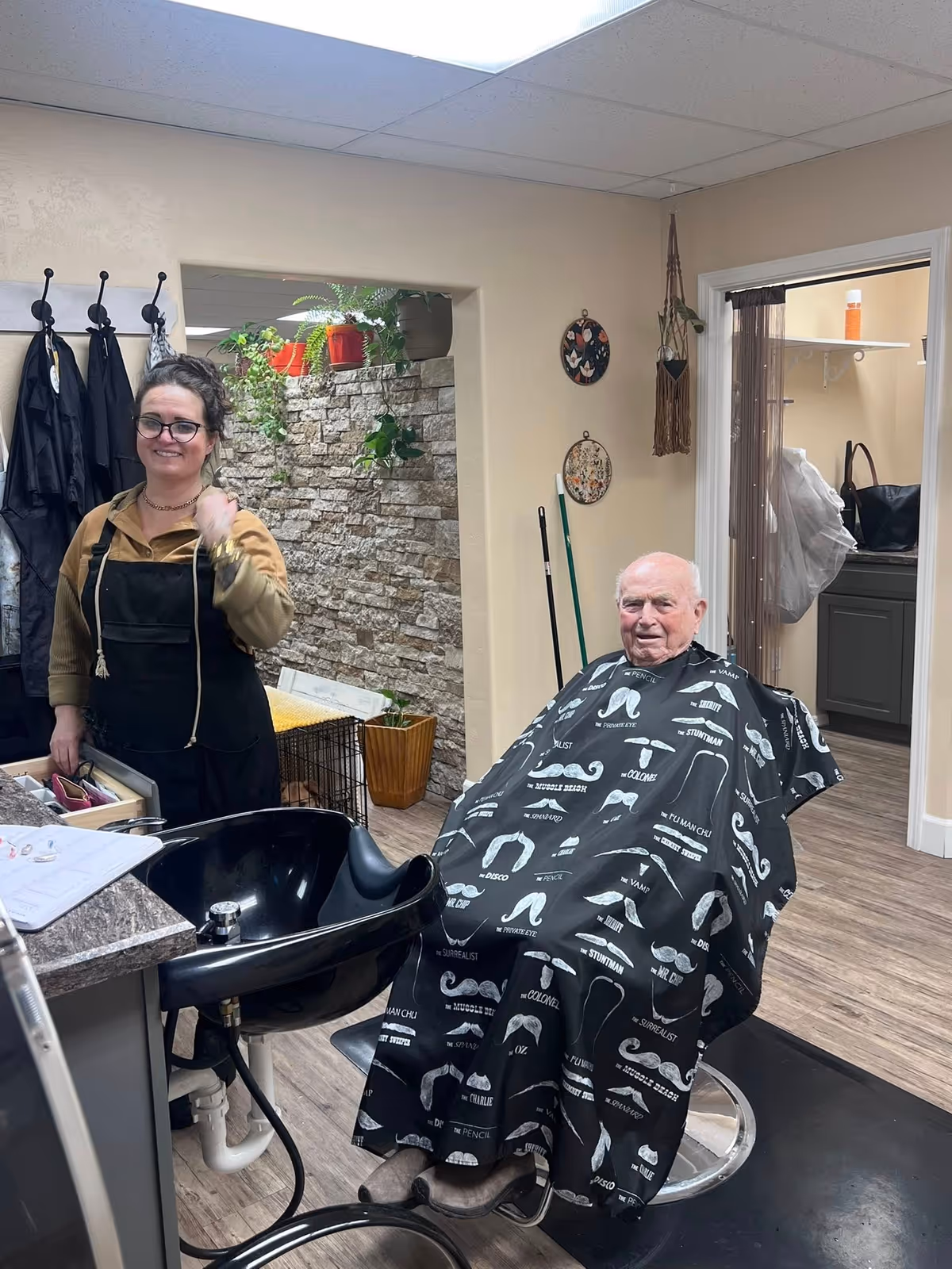 An elderly man is seated in a barber chair with a black haircut cape featuring various mustache styles printed on it. A woman wearing glasses and a black apron stands next to him, smiling. The room has light-colored walls, a stone accent wall with potted plants, and wooden flooring. There is a black wash basin in front of the man and some hairdressing tools on the counter.