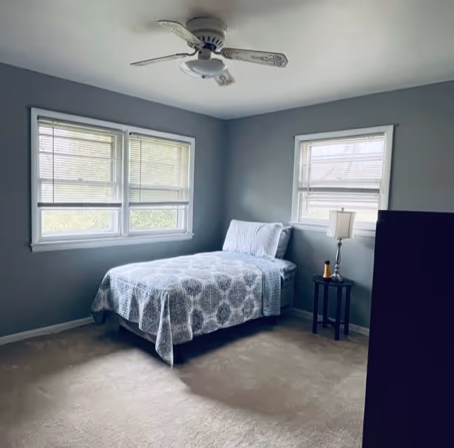 A simple bedroom with light blue walls and beige carpet. There is a single bed with a patterned blue and white bedspread and two pillows. The room has three windows with blinds partially open, letting in natural light. A small round side table next to the bed holds a lamp and a small object. A ceiling fan with a light fixture is mounted on the ceiling.