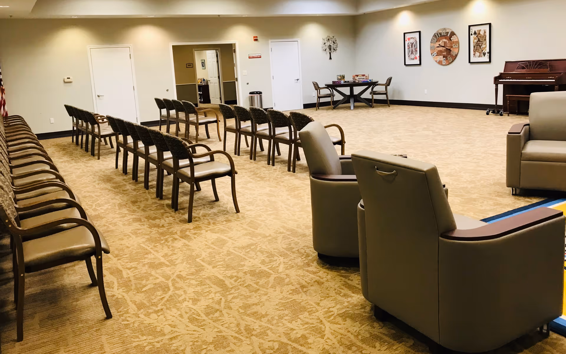 A large, well-lit room with rows of chairs arranged in a semi-circle facing an open space. The room has beige carpet and neutral-colored walls with framed artwork and a piano in the corner. There are two armchairs in the foreground and a table with chairs and board games in the background.